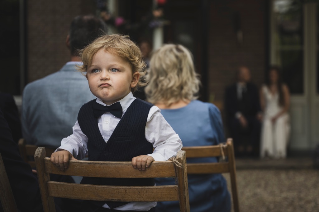 Jongetje met strik en gilet hangt over een stoel tijdens de trouwceremonie van Elena en Roeland, het bruidspaar onscherp op de achtergrond