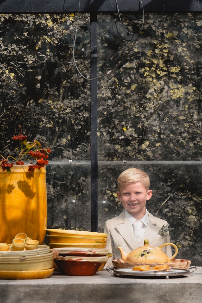 Jongen in licht kostuum lacht achter een tafel met geel aardewerk in de kas, bruiloft van Marlou en Freek op Domaine d’Heerstaayen